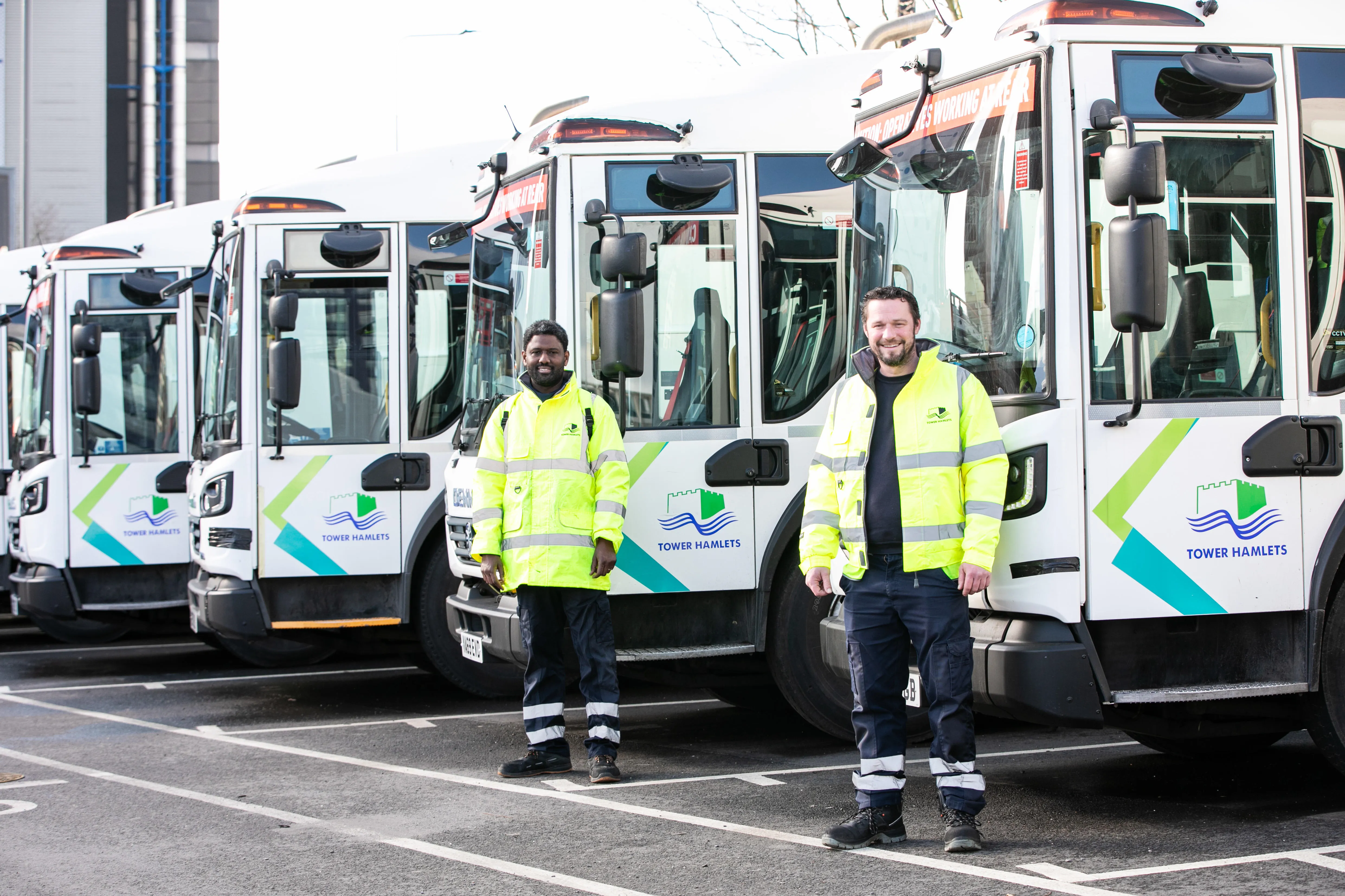 Two waste officers standing by four parked waste collection vehicles.