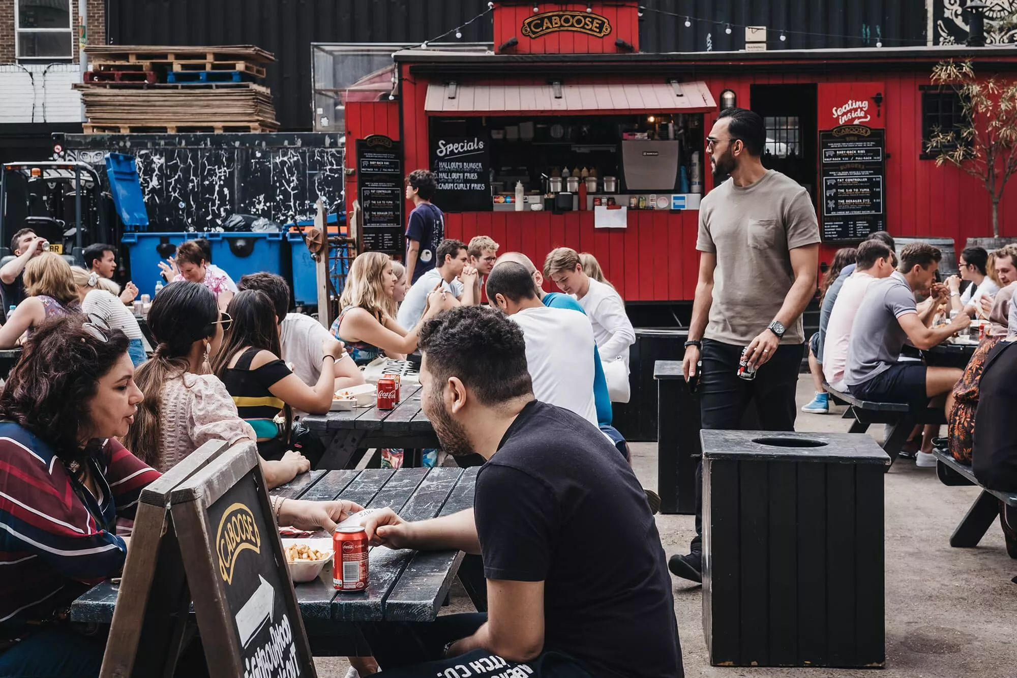 A street scene of people sitting at pub benches drinking and talking.